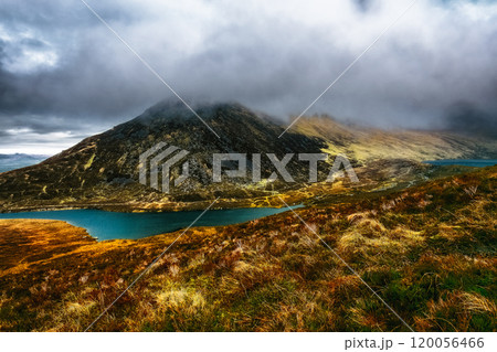A scenic view of a mountain range, with a foreground of dry grass, a curved lake in the valley, and a hilltop shrouded in mist. 120056466