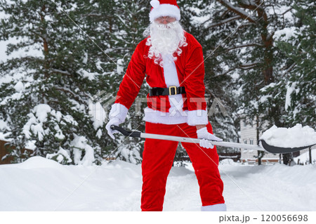 Santa Claus cleans snow with shovel in winter outdoors after a snowfall. Cleaning the streets in the village, clearing the passage for cars, difficult weather conditions for Christmas and New Year 120056698