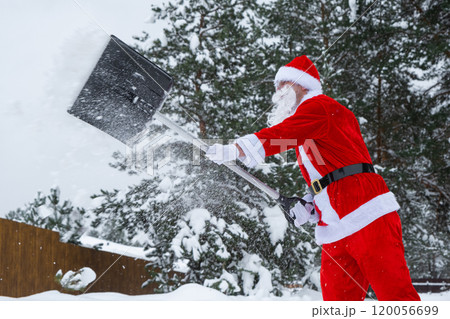 Santa Claus cleans snow with shovel in winter outdoors after a snowfall. Cleaning the streets in the village, clearing the passage for cars, difficult weather conditions for Christmas and New Year 120056699