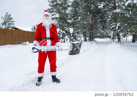 Santa Claus cleans snow with shovel in winter outdoors after a snowfall. Cleaning the streets in the village, clearing the passage for cars, difficult weather conditions for Christmas and New Year 120056702