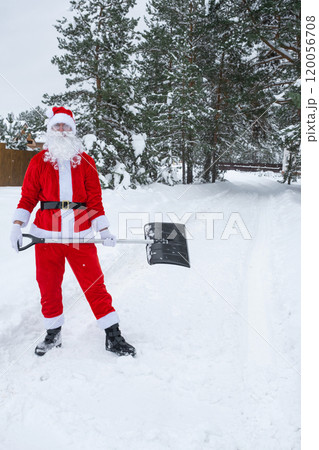 Santa Claus cleans snow with shovel in winter outdoors after a snowfall. Cleaning the streets in the village, clearing the passage for cars, difficult weather conditions for Christmas and New Year Santa Claus cleans snow with shovel in winter outdoors after a snowfall. Cleaning the streets in the village, clearing the passage for cars, difficult weather conditions for Christmas and New Year 120056708