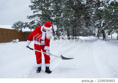 Santa Claus cleans snow with shovel in winter outdoors after a snowfall. Cleaning the streets in the village, clearing the passage for cars, difficult weather conditions for Christmas and New Year 120056903