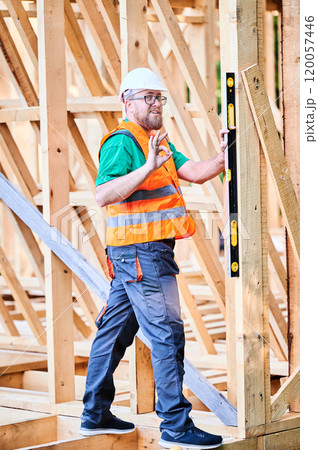 Carpenter constructing wooden frame house. Man inspects walls for levelness using spirit level, showing gesture OK sign, wearing protective overalls, helmet and orange vest. 120057446