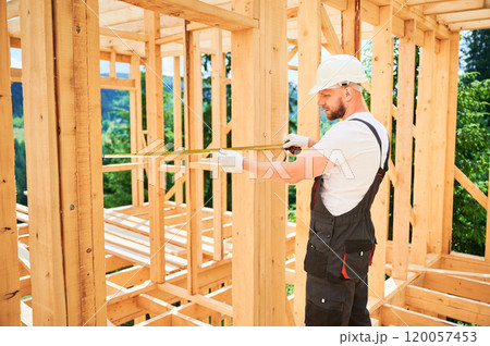Carpenter building wooden frame house. Man measures the distance with tape measure while dressed in workwear and a helmet. Carpenter building wooden frame house. Man measures the distance with tape measure while dressed in workwear and a helmet. 120057453