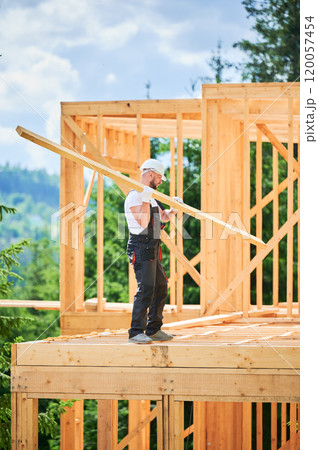Carpenter builds wooden frame house near woods. Bearded man holds large board on shoulder, wearing work clothes and helmet. Concept of modern and sustainable building. Carpenter builds wooden frame house near woods. Bearded man holds large board on shoulder, wearing work clothes and helmet. Concept of modern and sustainable building. 120057454