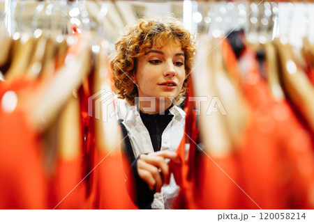 young attractive red-haired woman choosing red clothes in boutique 120058214