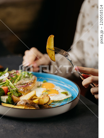 Close-up of hand holding a roasted potato slice over a breakfast plate with eggs and greens 120058514