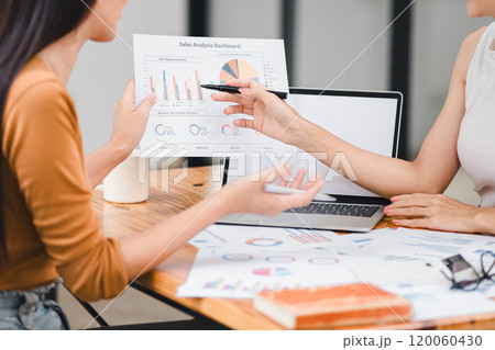 Two professionals analyze business data using laptop displaying sales dashboard and various printed charts on desk. 120060430
