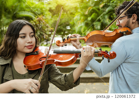 Violinist man and woman back to back playing violin in a park outdoors. Two young violinists standing playing violin in a park. Portrait of man and woman together playing violin in park Violinist man and woman back to back playing violin in a park outdoors. Two young violinists standing playing violin in a park. Portrait of man and woman together playing violin in park 120061372