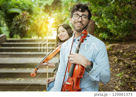 Portrait of two young violists sitting outside. Man and woman violinist sitting on the stairs. Concept of two young violinists, Two young violinists sitting outdoors looking at the camera Portrait of two young violists sitting outside. Man and woman violinist sitting on the stairs. Concept of two young violinists, Two young violinists sitting outdoors looking at the camera 120061373