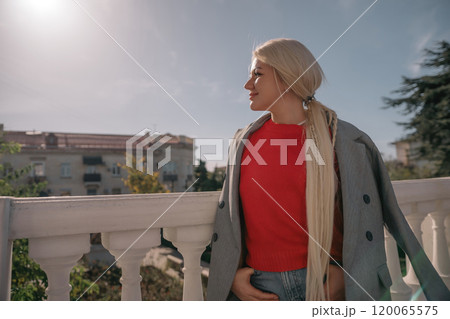 A blonde woman wearing a red shirt and gray jacket stands on a balcony. She is wearing ripped jeans and has her hands in her pockets. A blonde woman wearing a red shirt and gray jacket stands on a balcony. She is wearing ripped jeans and has her hands in her pockets. 120065575