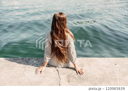 A woman is sitting on a ledge by a body of water, wearing a dress with a leopard print pattern. The scene is peaceful and serene, with the woman enjoying the view of the water and the surrounding area 120065639