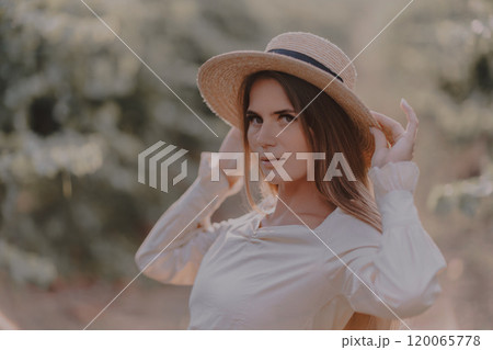 Woman with straw hat stands in front of vineyard. She is wearing a light dress and posing for a photo. Travel concept to different countries Woman with straw hat stands in front of vineyard. She is wearing a light dress and posing for a photo. Travel concept to different countries 120065778