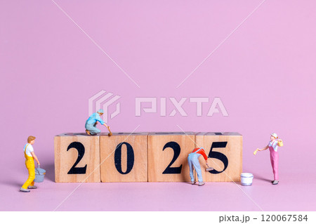Miniature figures in overalls paint and construct the numbers "2025" on wooden blocks 120067584