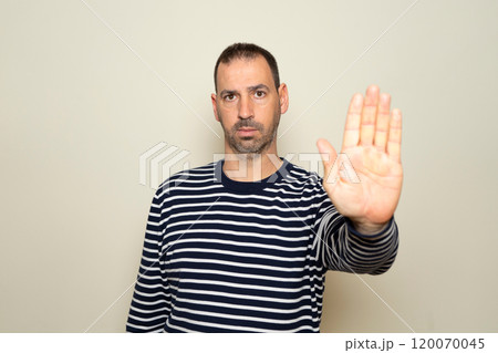 Portrait of a bearded Hispanic man in his 40s wearing a striped sweater and making a stop gesture with his palm, rejecting stress and problems, without discrimination. Isolated on beige background Portrait of a bearded Hispanic man in his 40s wearing a striped sweater and making a stop gesture with his palm, rejecting stress and problems, without discrimination. Isolated on beige background 120070045