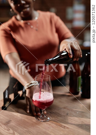 Vertical shot of female African American sommelier pouring craft cider into glass during product sampling in tasting room as part of factory, copy space 120071362