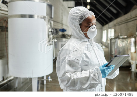 Medium shot of professional African American female technician in hazmat suit using tablet computer inspecting stainless tanks while controlling production process at food factory, copy space 120071489