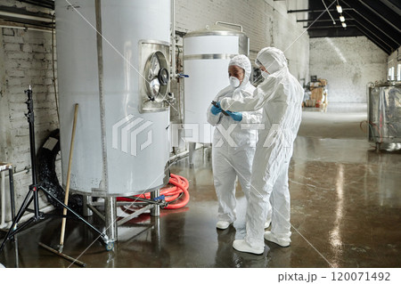 Full length shot of two professional female supervisors in masks and coveralls using tablet computer discussing production process while inspecting tanks in factory workshop Full length shot of two professional female supervisors in masks and coveralls using tablet computer discussing production process while inspecting tanks in factory workshop 120071492
