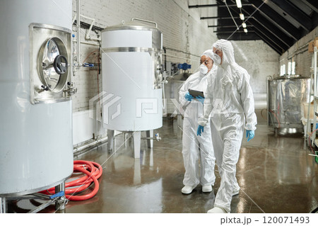 Full length shot of professional female supervisor in mask and coverall walking towards stainless tank with liquid while inspecting workshop equipment with colleague at factory 120071493