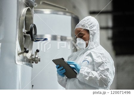 Medium shot of female African American technician in mask and hazmat suit using tablet computer inspecting stainless tanks while controlling fermentation process at cider factory, copy space Medium shot of female African American technician in mask and hazmat suit using tablet computer inspecting stainless tanks while controlling fermentation process at cider factory, copy space 120071496