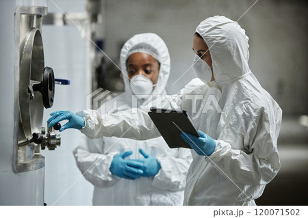 Side view of female process technician in hazmat suit using tablet computer inspecting stainless tanks with colleague while controlling fermentation process at craft cider factory, copy space Side view of female process technician in hazmat suit using tablet computer inspecting stainless tanks with colleague while controlling fermentation process at craft cider factory, copy space 120071502