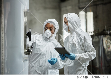 Two professional female process technicians wearing masks and hazmat suits inspecting equipment in cider factory workshop while collaboratively controlling product production, copy space 120071504