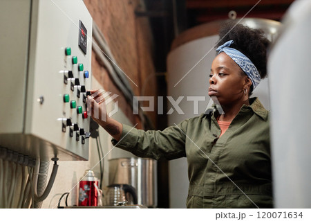 Side view of focused African American female production worker operating machine with panel buttons while looking at indicators at food factory, copy space 120071634
