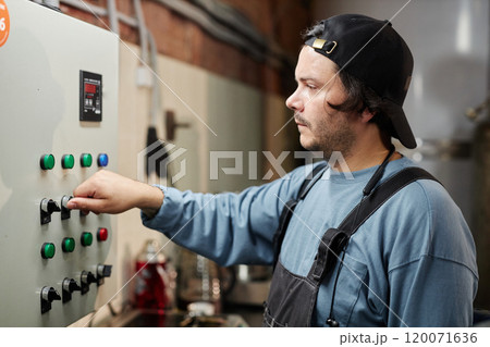 Side view of concentrated male production worker operating factory machine adjusting panel buttons while looking at indicators at food plant, copy space 120071636
