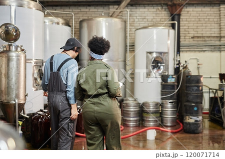 Rear view of diverse team of two cider factory workers inspecting production process on workshop floor with huge fermentation tanks at plant, copy space 120071714
