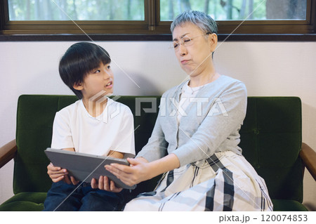 家でタブレットを一緒に楽しむ孫と老眼でよく見えない祖母 120074853
