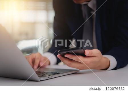 Business professional using a smartphone while working on a laptop. Suitable for modern business communication, remote work, or digital technology Business professional using a smartphone while working on a laptop. Suitable for modern business communication, remote work, or digital technology 120076582