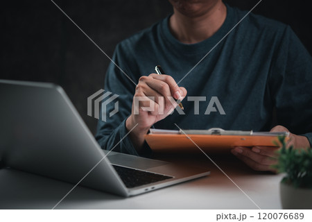 Businessperson writing on a clipboard while working on a laptop, representing planning and organization. Suitable for project management content Businessperson writing on a clipboard while working on a laptop, representing planning and organization. Suitable for project management content 120076689