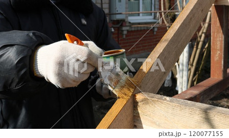 gloved hands holding a paintbrush and tin can in front of a wooden outbuilding frame structure to apply a protective coat of paint or varnish to the wood gloved hands holding a paintbrush and tin can in front of a wooden outbuilding frame structure to apply a protective coat of paint or varnish to the wood 120077155