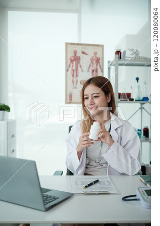 A woman in a white lab coat is sitting at a desk with a laptop 120080064