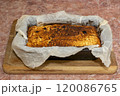 A baked cheesecake with a golden brown crust lies in a black pan on a wooden cutting board in a cozy kitchen setting. 120086765