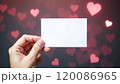 A hand holds a blank white card in front of a blurred background with red heart shapes. Concept of love and romance. Valentine's day background 120086965