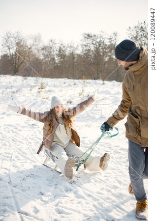 Portrait of romantic couple having fun on winter walk. Man carry his wife on a sled. Family wearing warm jacket and knitted hats. 120089447