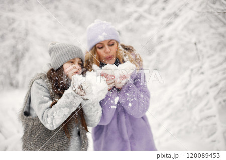 Young blonde woman with her daughter playing in winter in a snowballs. Caucasian woman wearing purple warm jacket and knitted hat. Brunette girl wearing grey jacket and hat. 120089453