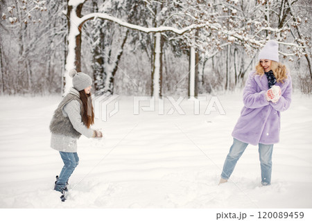 Young blonde woman with her daughter playing in winter in a snowballs. Caucasian woman wearing purple warm jacket and knitted hat. Brunette girl wearing grey jacket and hat. 120089459