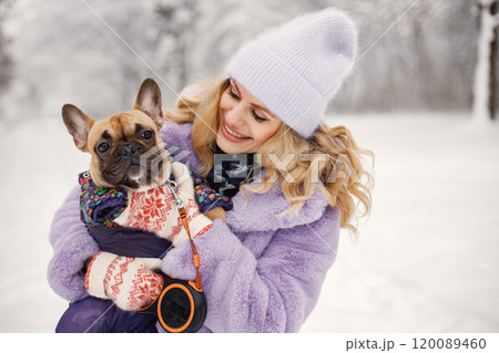 Young blonde woman with her dog french bulldog playing in winter on a snow. Caucasian woman wearing purple warm jacket and knitted hat. Dog wearing warm overalls. Young blonde woman with her dog french bulldog playing in winter on a snow. Caucasian woman wearing purple warm jacket and knitted hat. Dog wearing warm overalls. 120089460