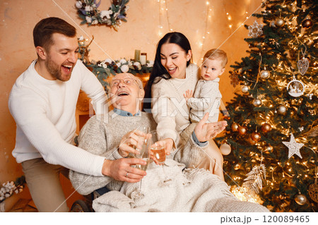 Young family celebrating Christmas together. Brunette mother, bearded father, blonde son and old grandfather on a wheelchair sitting near Christmas tree and drinking champagne. Family wearing beige Young family celebrating Christmas together. Brunette mother, bearded father, blonde son and old grandfather on a wheelchair sitting near Christmas tree and drinking champagne. Family wearing beige 120089465