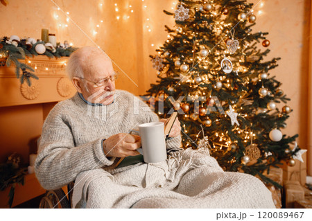 Old grey man celebrating Christmas alone. Man on a wheelchair sitting near Christmas tree. Grandfather covering on blanket, drinking tea and reading a book. 120089467