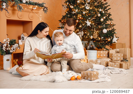 Young family celebrating Christmas together. Brunette mother, bearded father and blonde son sitting near Christmas tree and reading a book. Family wearing beige knitted clothes. 120089487