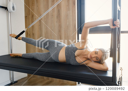 Pregnant woman doing exercises with a special equipment. Fit blonde woman working out in a gym. Woman wearing grey sportive leggins and top. 120089492
