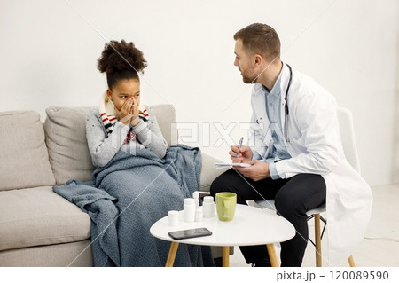 Male doctor in white medical gown asking sick girl about her symptoms. Black girl has fever and overspread blanket. Different medicines are on table near sofa. 120089590