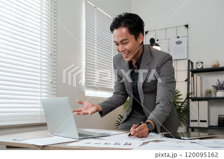 A man in a suit is sitting at a desk with a laptop open and a smile on his face 120095162