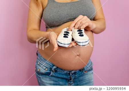 A joyful expectant mother holding tiny baby sneakers against her pregnant belly 120095934