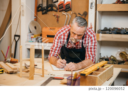 Man in plaid shirt and overalls working on a wooden project in his workshop, drawing on clipboard with pencil. Surrounded by tools and wood, woodworking and DIY craftsmanship, National Carpenters Day Man in plaid shirt and overalls working on a wooden project in his workshop, drawing on clipboard with pencil. Surrounded by tools and wood, woodworking and DIY craftsmanship, National Carpenters Day 120096052