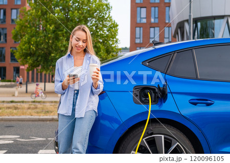 Smiling woman holding a cup of coffee checks the battery level and makes a payment through an app for charging her EV on the street. Smiling woman holding a cup of coffee checks the battery level and makes a payment through an app for charging her EV on the street. 120096105