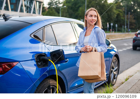 Woman parks her electric car and begins charging it while carrying shopping bags in the evening Woman parks her electric car and begins charging it while carrying shopping bags in the evening 120096109